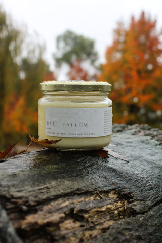 A jar of Highlands Natural Beef Tallow with the label showing the product name, rendered from grass-fed Australian beef by Highlands Natural. The jar is sealed with a gold lid and sits on a log outside in the rain with orange leaves.