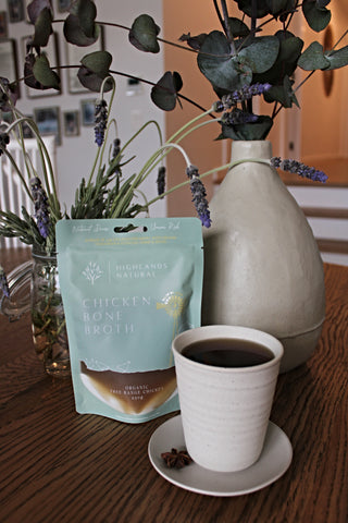 Pouch of Organic Free-Range Chicken Bone Broth by Highlands Natural with a ceramic cup filled with broth and star anise on the saucer. Two vases with lavender and gum leaves, sitting on a wooden table.